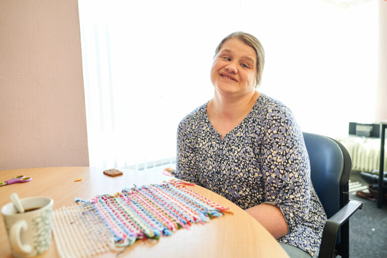 Jonge vrouw zit aan tafel te borduren met een kopje koffie en koekje erbij