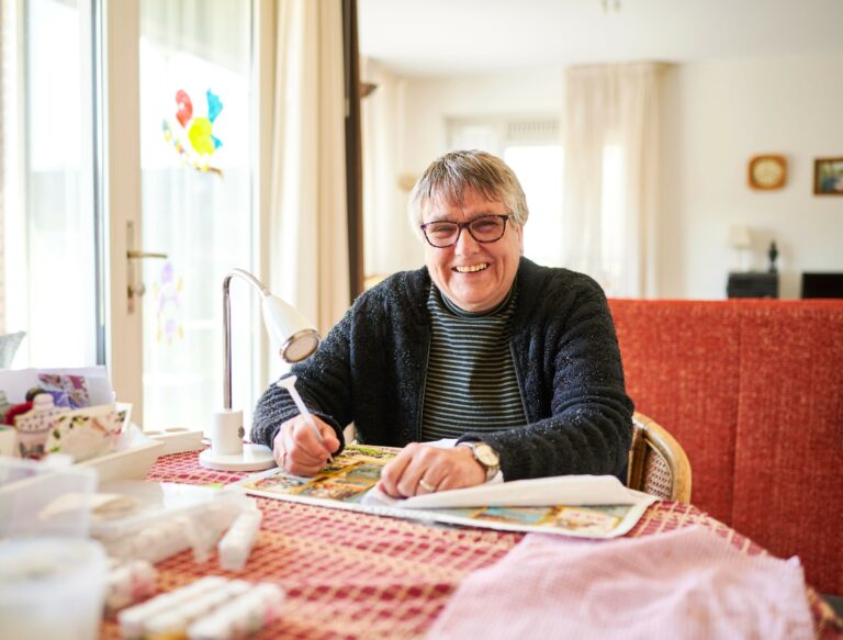 Oudere lachende vrouw zit aan tafel een diamond painting schilderij te maken