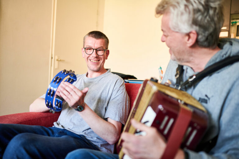 Twee mannen zitten naast elkaar muziek te maken. 1 met een accordeon en de andere met een ...