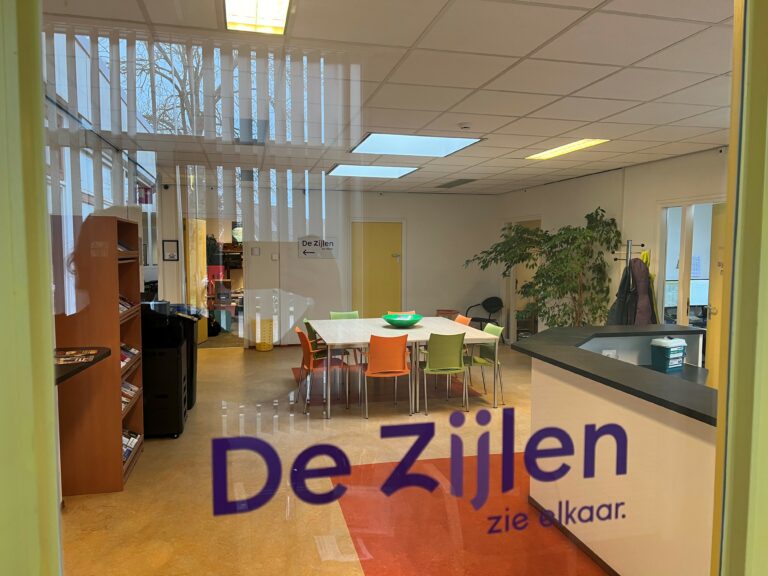 Ruimte met een kast en een grote tafel met gekleurde stoelen eromheen Tekst op raam De Zijlen zie elkaar