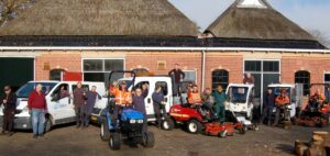 Groep mannen in werkkleding voor een boerderij, tussen werkbussen en zitmaaiers