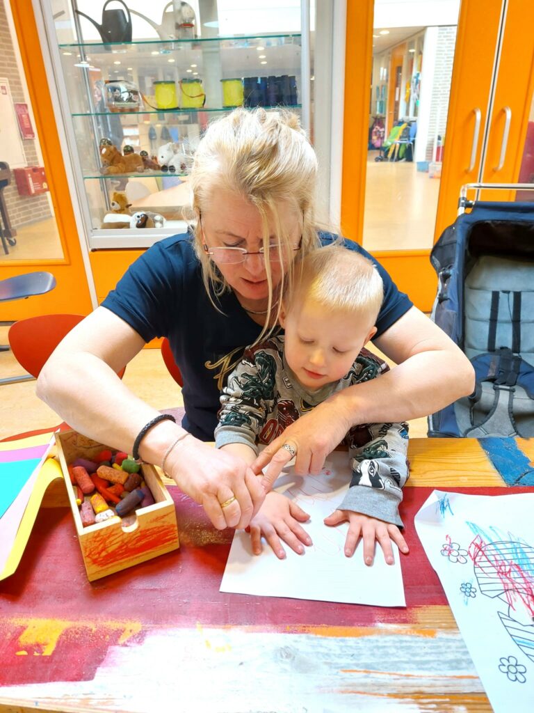 Vrouw met lang blond haar zit achter klein jongetje om zijn handen te tekenen