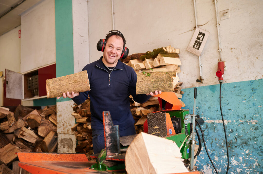 Vrolijk lachende jonge man met gehoorbescherming tussen stukken hout en een kloofmachine