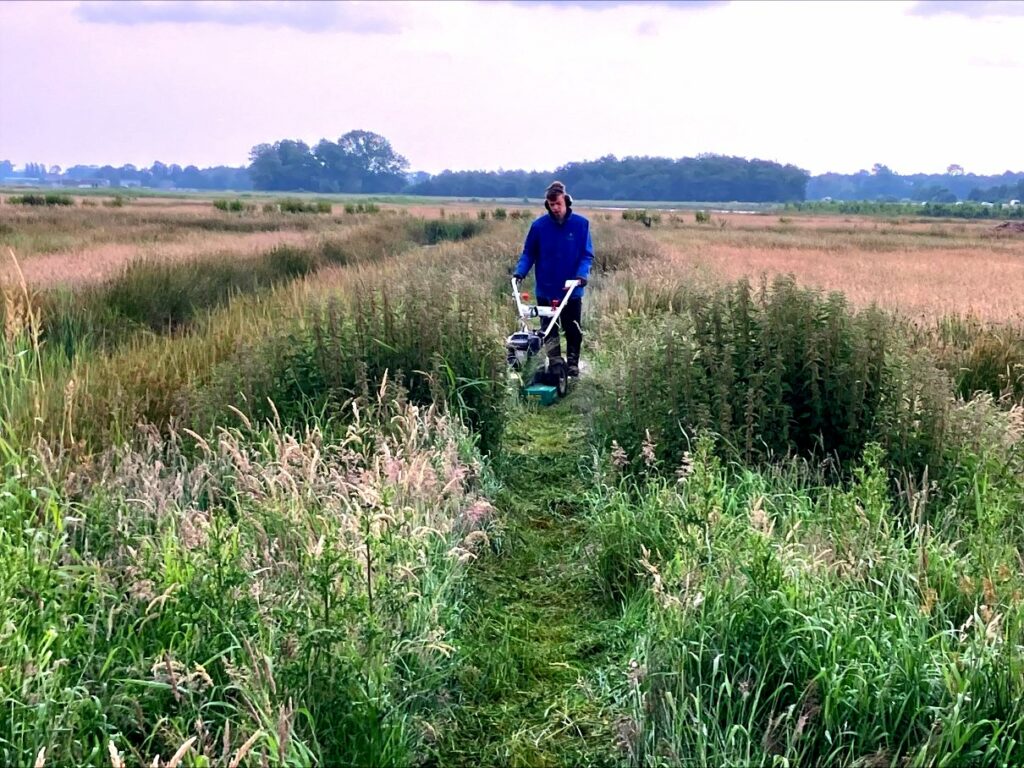 Jonge man in blauwe jas op groen heideveld die achter een machine loopt