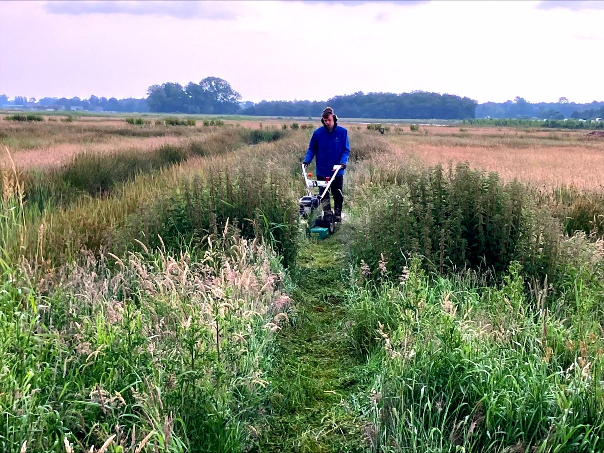 Jonge man in blauwe jas op groen heideveld die achter een machine loopt