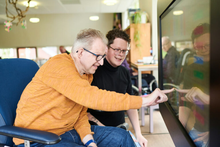 Oudere man zit met een jonge man voor beeldtelevisie en wijst iets aan