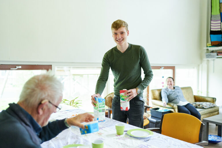 Jonge man zet melk en karnemelk op tafel voor oudere man die boter in zijn hand heeft vrouw kijkt toe vanaf de bank