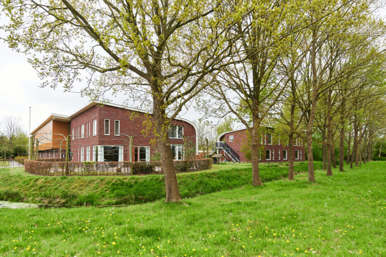 Drie gebouwen met sloot eromheen groen gras en rij bomen