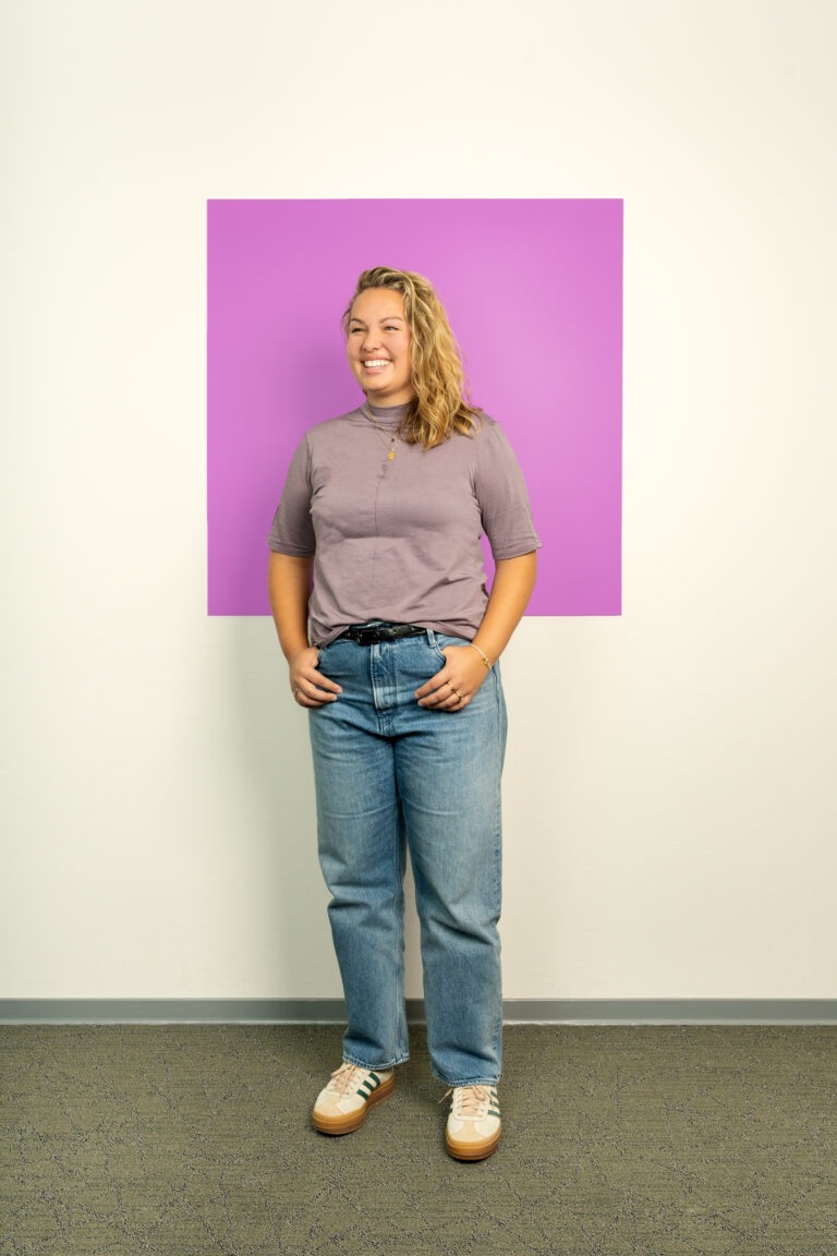 Lachende jonge vouw met lang krullend haar staat met handen in haar zakken voor lila vierkant