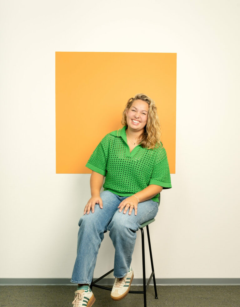 Lachende jonge vouw met lang krullend haar en groene trui zit op stoel voor licht oranje vierkant