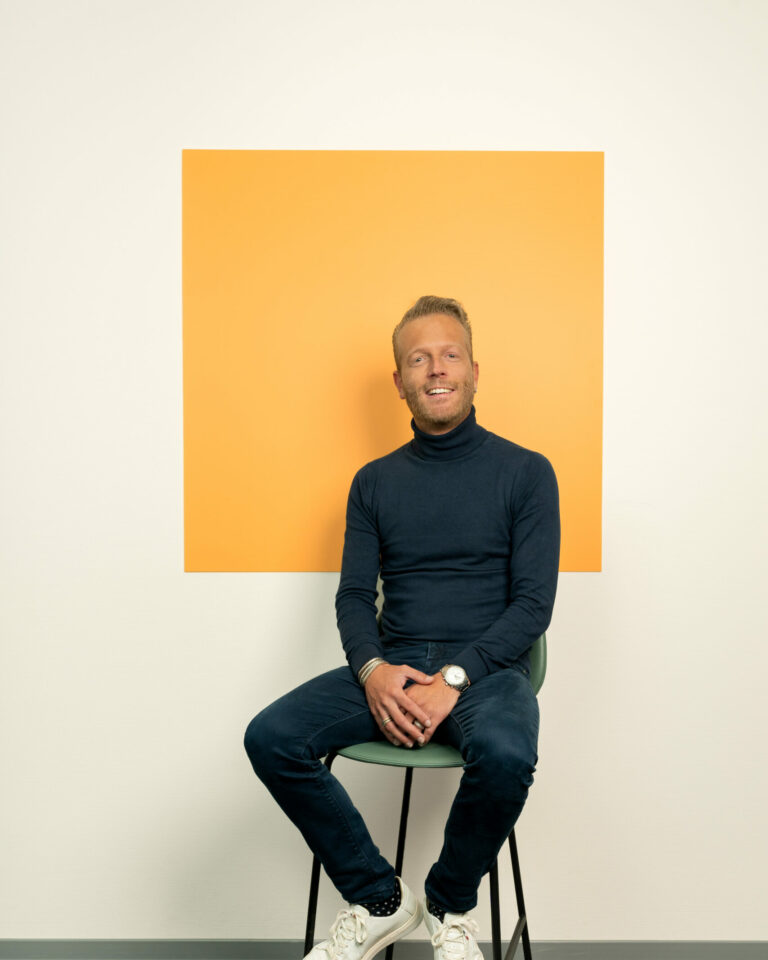 Man met korte baard lachend zit op stoel voor licht oranje vierkant