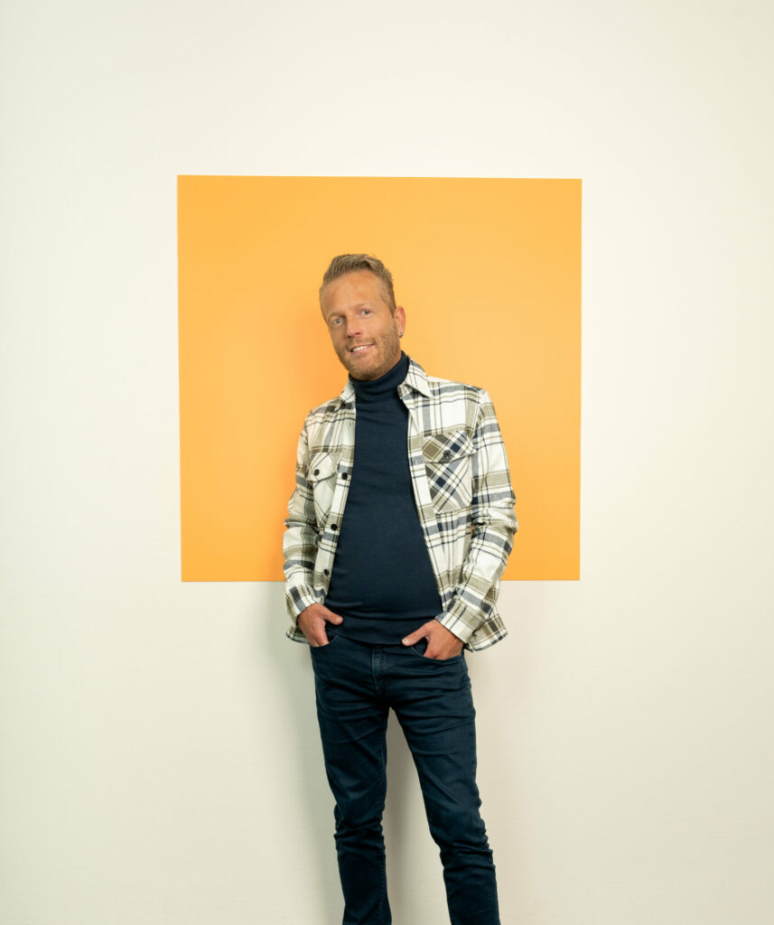 Jonge man met korte baard en geblokt overhemd staat voor licht oranje vierkant
