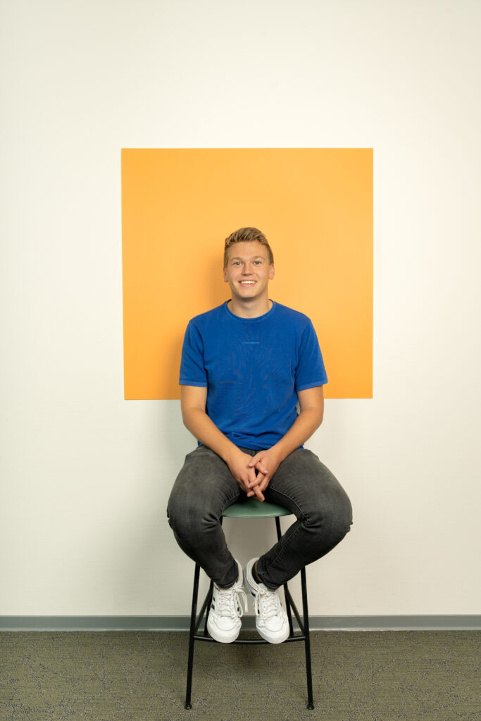 Jonge man in blauw tshirt zit op kruk voor een licht oranje vierkant
