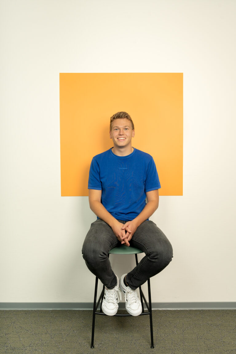 Jonge man in blauw tshirt zit op kruk voor een licht oranje vierkant