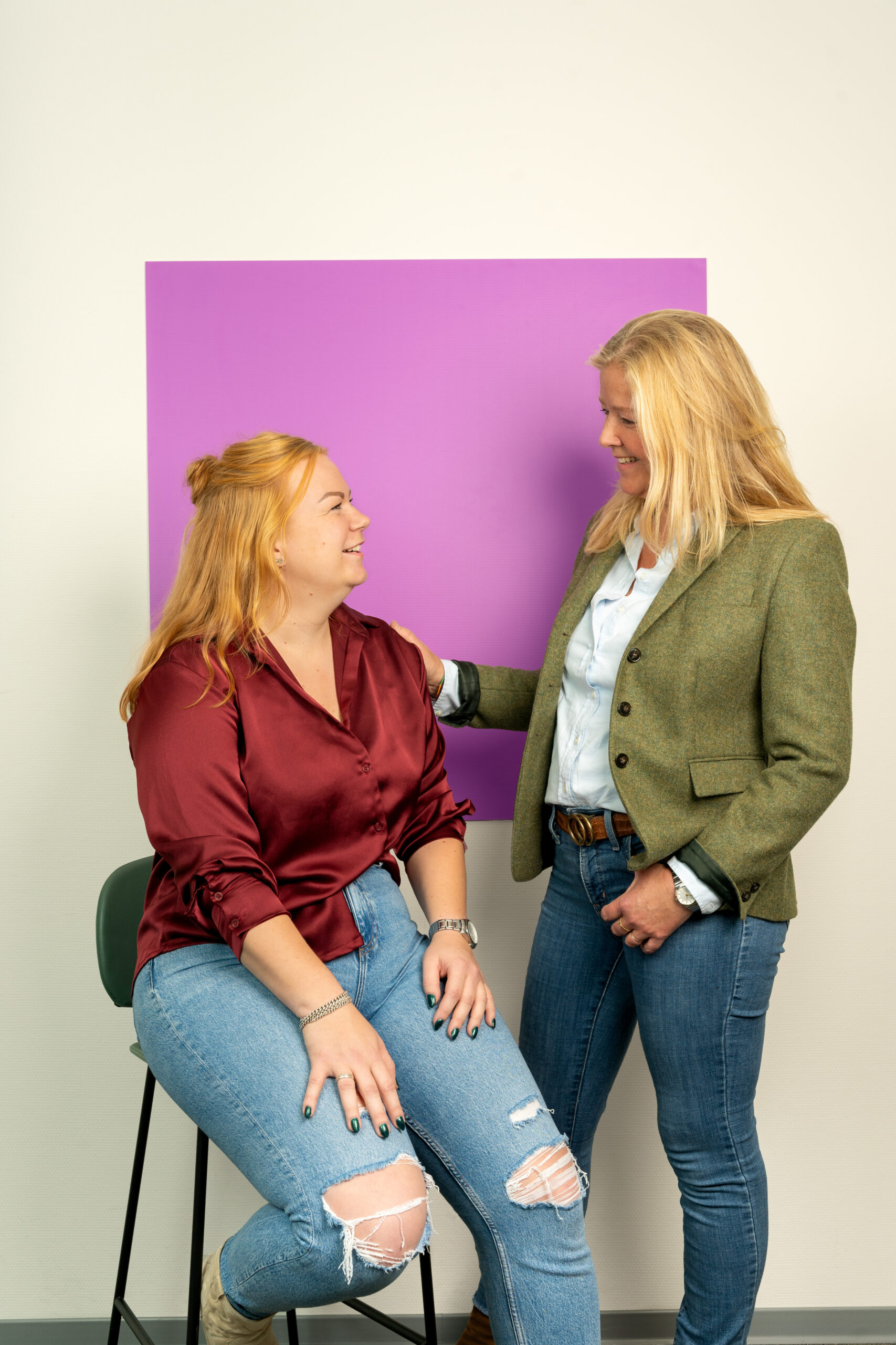 Twee vrouwen met lang blond haar. 1 heeft hand op de schouder van degene die zit