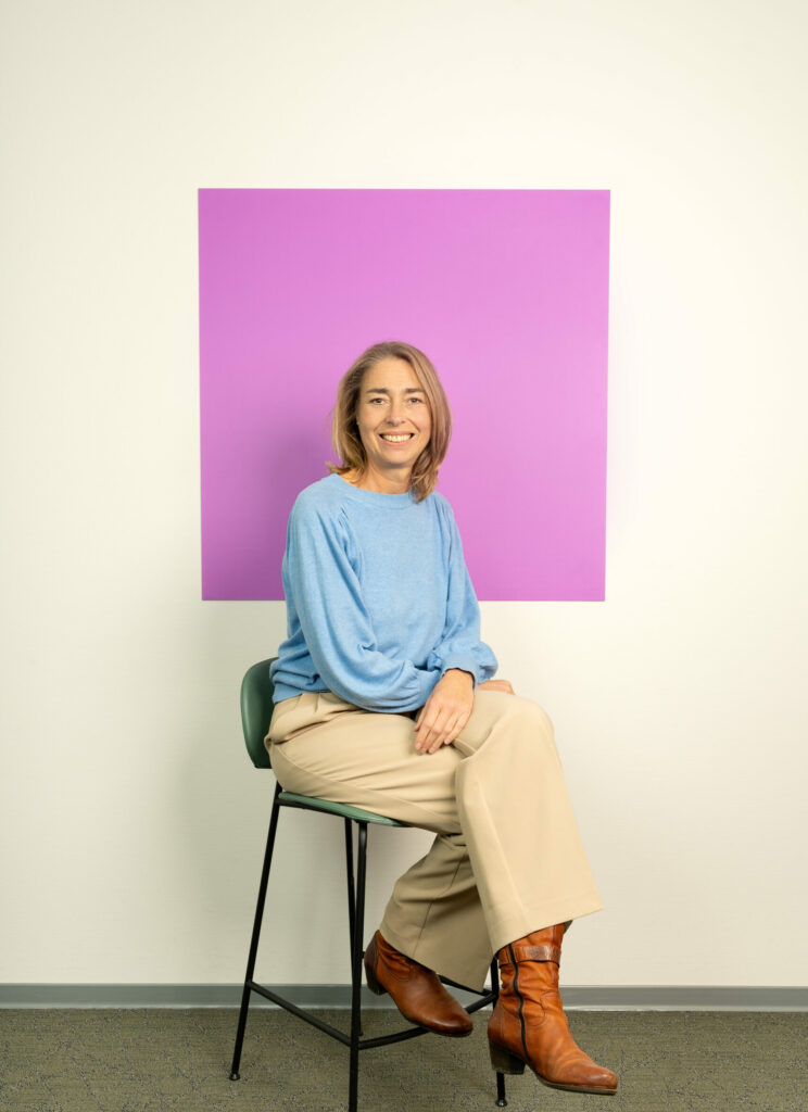 Lachende vrouw met halflang bruinhaar in blauwe trui zit voor lila vierkant