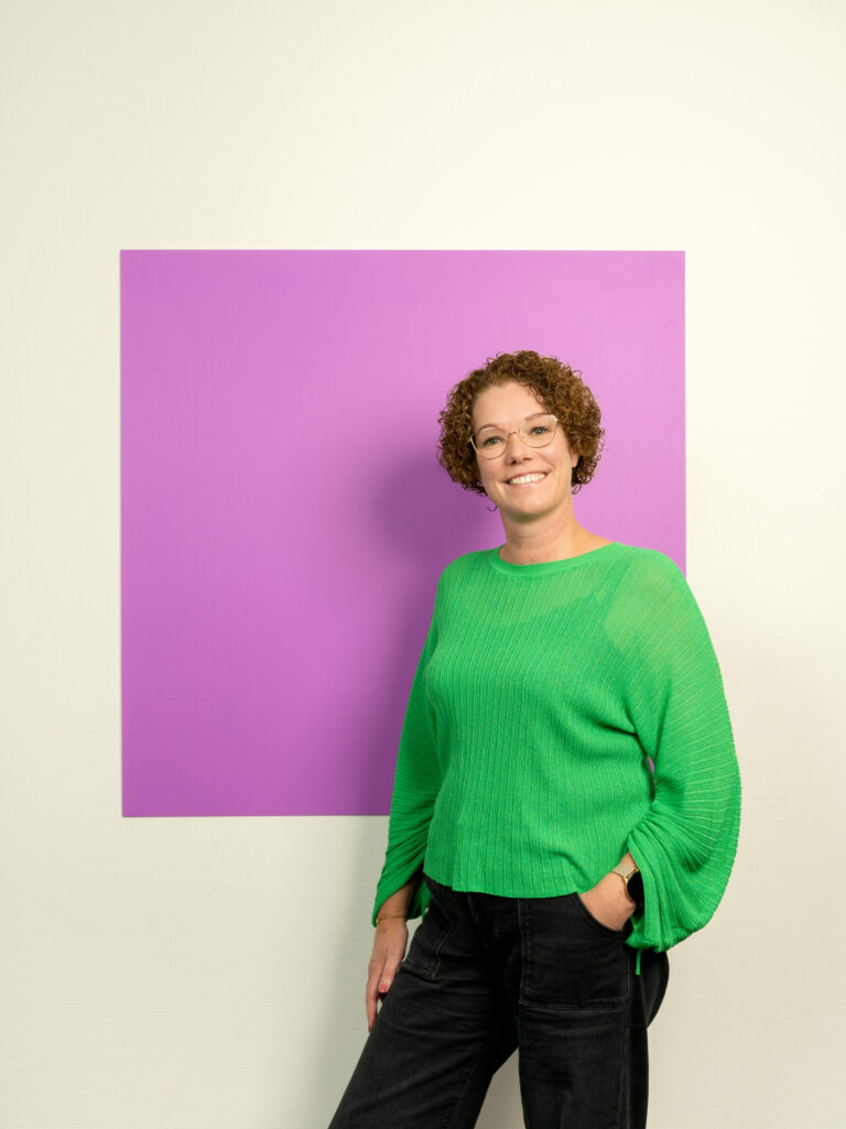 Jonge vrouw met krullen haar in groene trui en zwarte broek staat voor lila vierkant