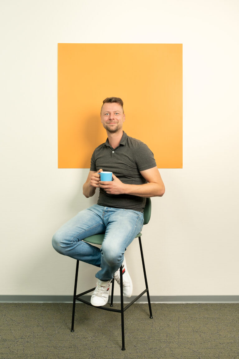 Man met blauw koffie kopje in zijn hand zit op een kruk voor een oranje/geel vierkant
