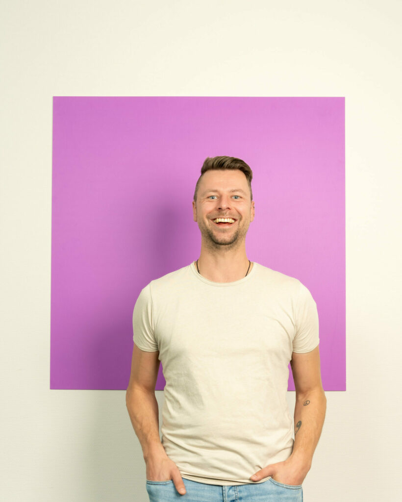 Jonge man in licht t-shirt staat lachend voor een roze/lila vierkant
