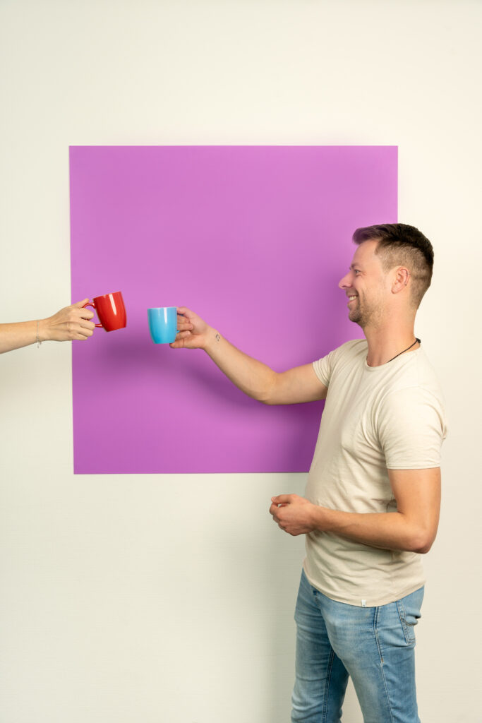 Jonge man proost met blauw koffiekopje staand voor een roze/lila vierkant