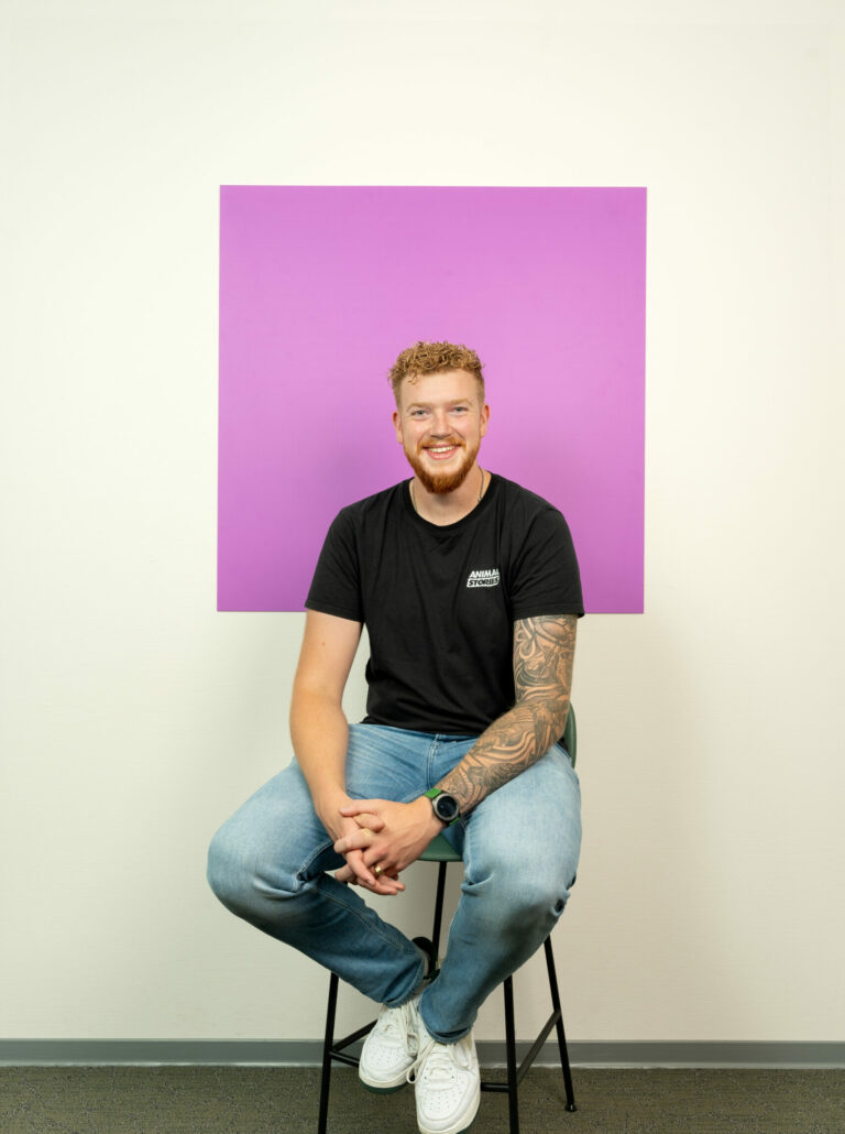 Jonge man met tatoeages in spijkerbroek zit op een kruk voor een paars/roze vierkant