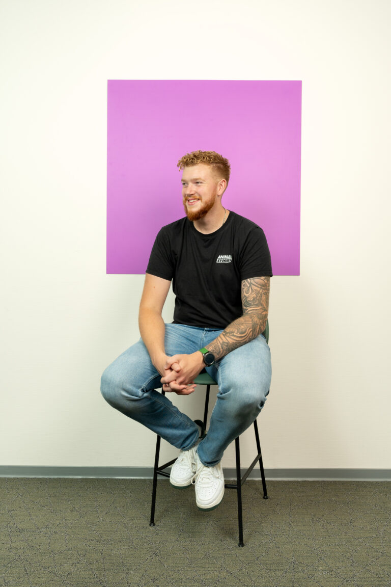 Jonge man met tatoeages in spijkerbroek zit op een kruk voor een paars/roze vierkant
