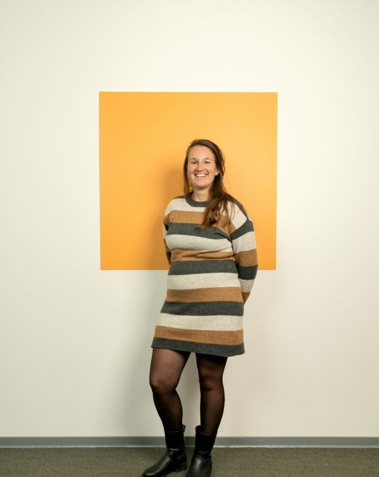 Jonge vrouw met lang bruin haar in gestreepte jurk staat voor oranje/geel vierkant