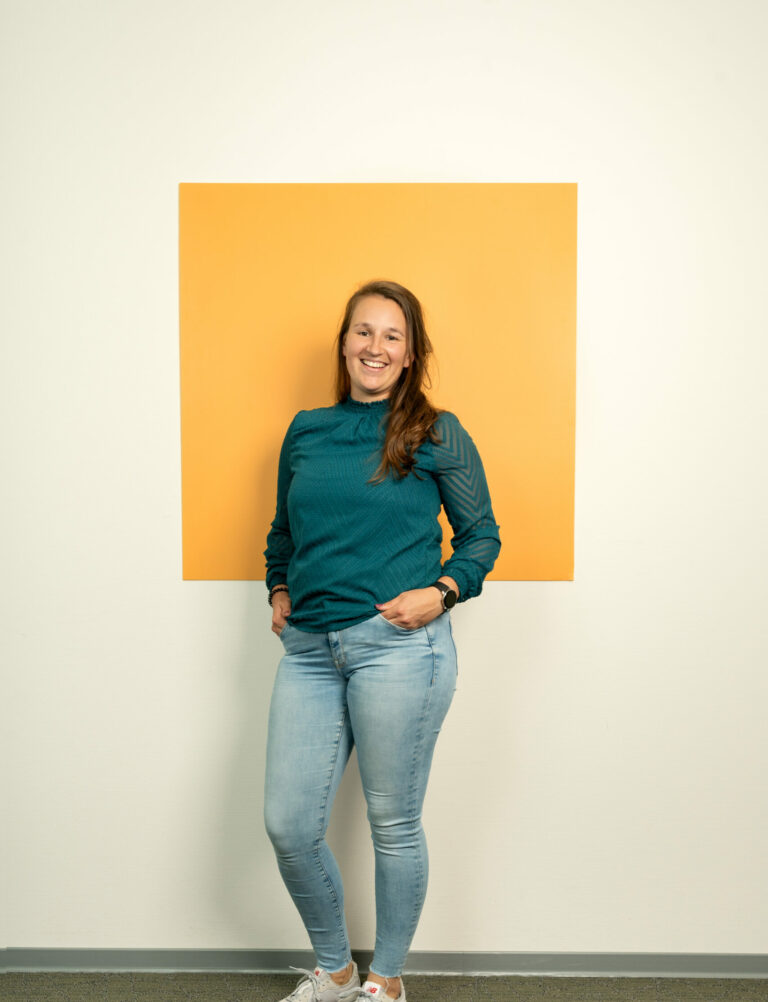 Jonge vrouw met lang bruin haar in spijkerbroek staat voor oranje/geel vierkant