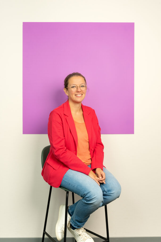Vrouw in rode blazer en spijkerbroek zit op een kruk voor een lila vierkant