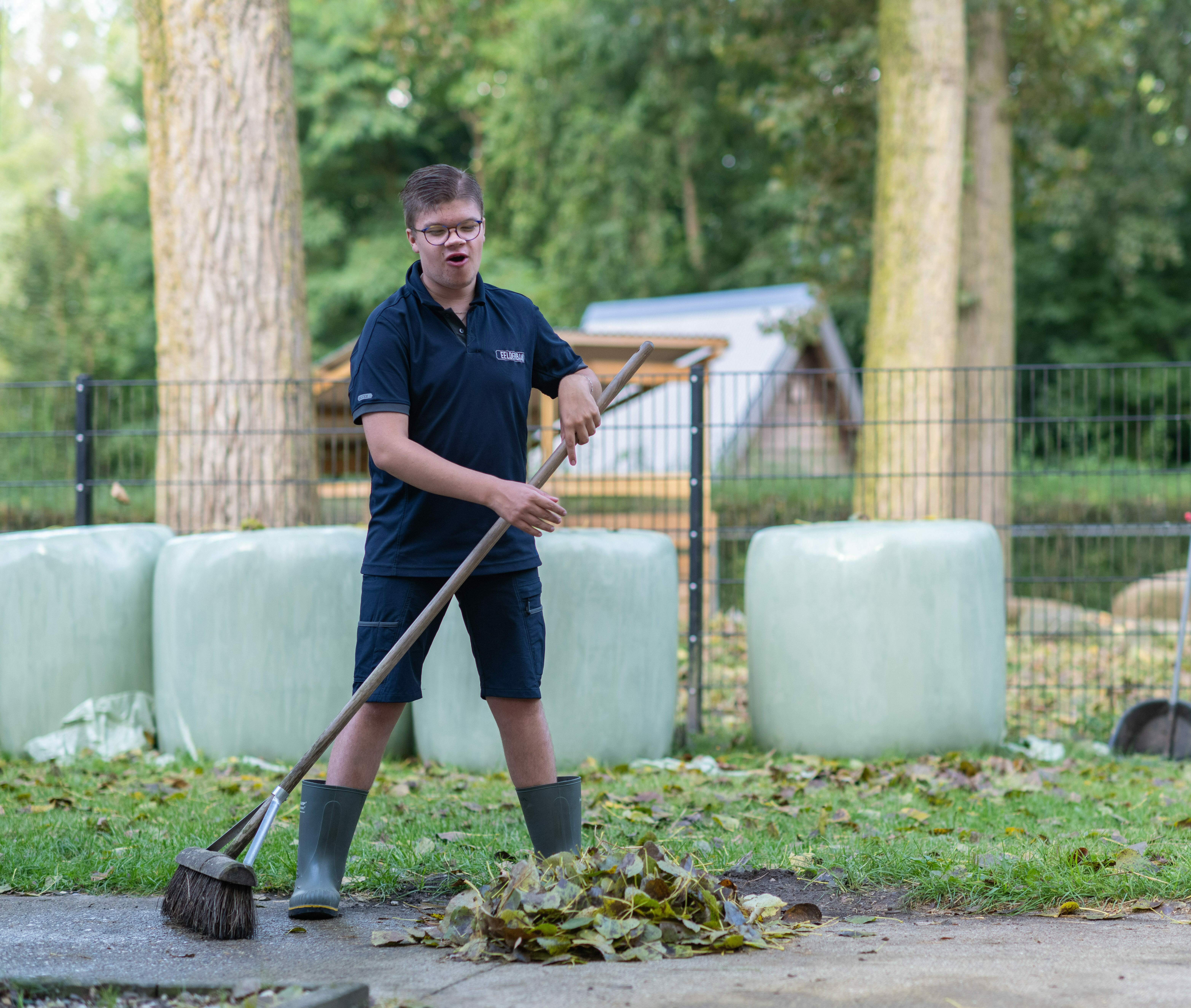 Jonge man in donkerblauwe lirte broek en tshirt veegt met veger bladeren bij elkaar op een dierenweide