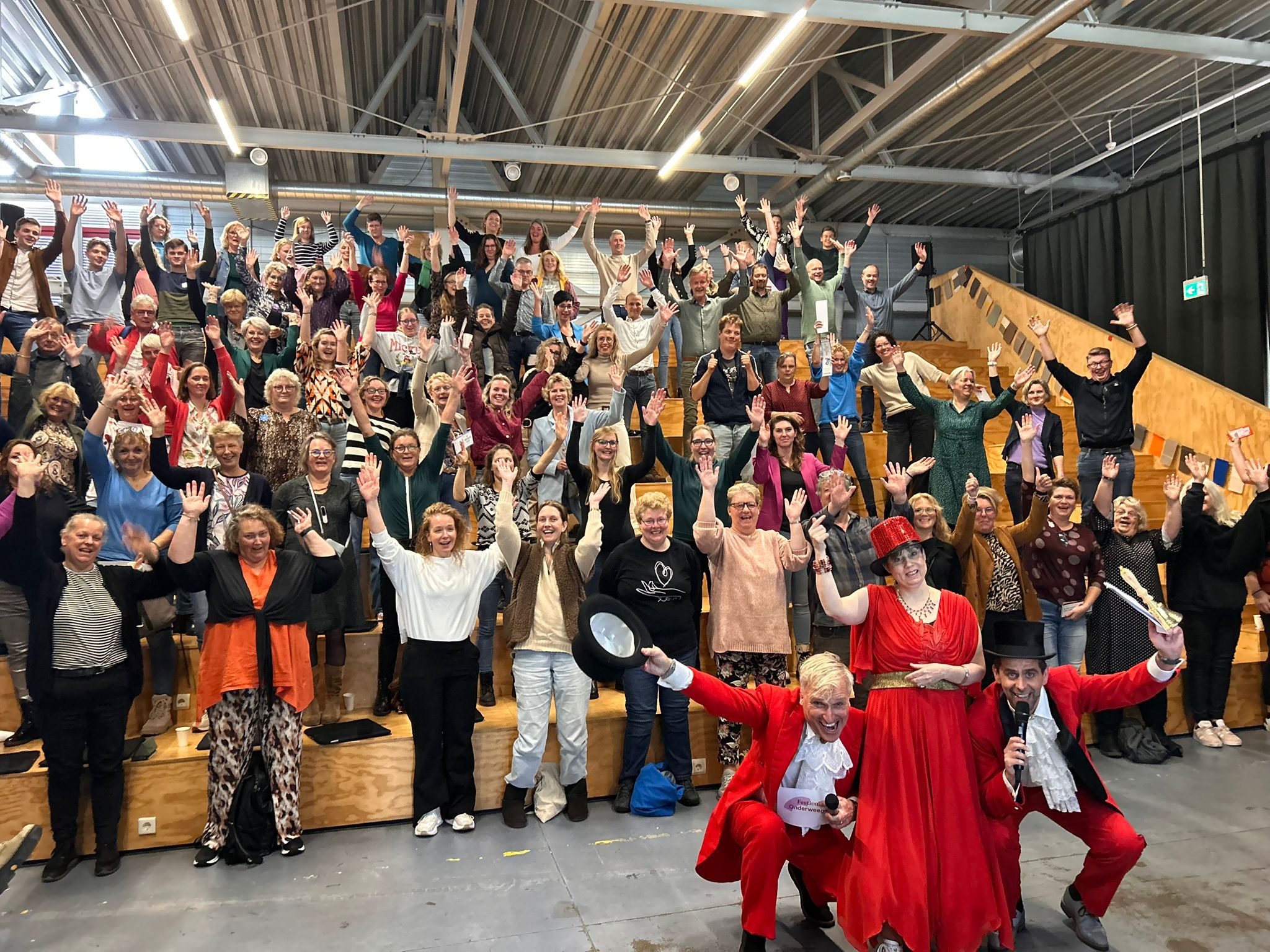 Heel veel mensen staan op een podium met armen omhoog. Op voorgrond 2 mannen en een vrouw in mooie rode kleding