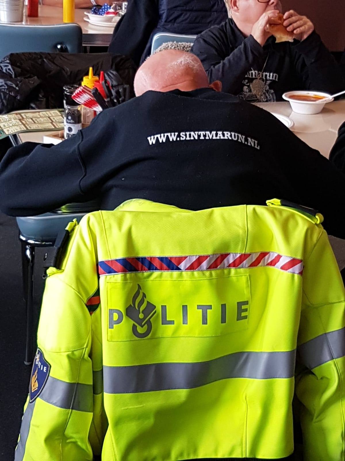 Man aan een tafel die broodje en soep eet en een man waarvan je de rug ziet en een politiejas over de stoel