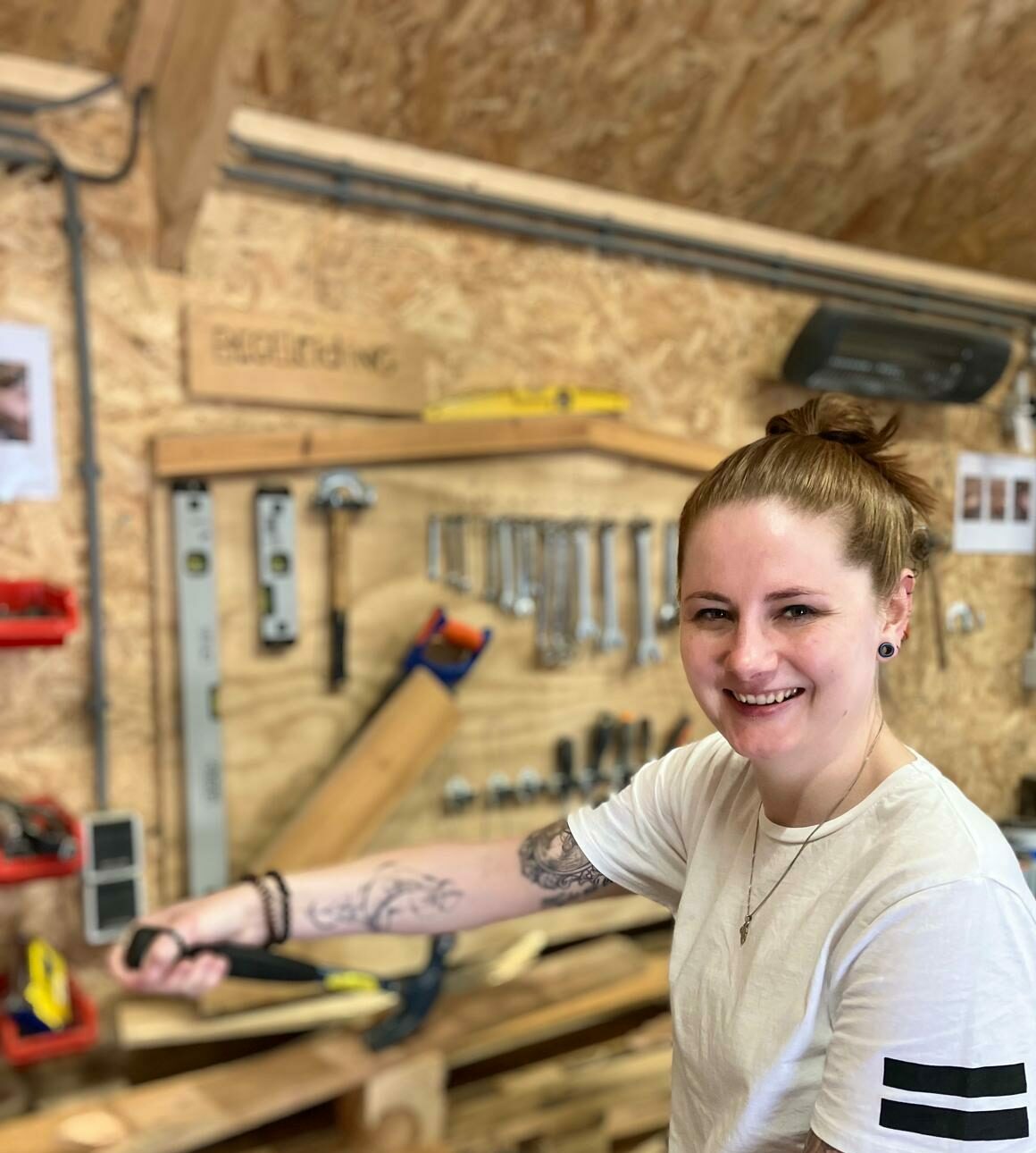 Vrouw met knotje in haar haar en tatoages op haar arm staat achter een werkbank met gereedschap