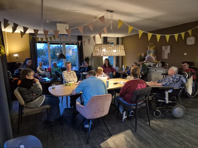 Volle huiskamer met personen om een tafel. Kamer is versierd met vlaggetjes